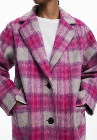 Cappotto in lana a scacchi rosa e grigio con colletto a revers, due tasche frontali e bottoni neri. Texture morbida e motivo a quadri evidente.