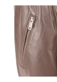 Braune Lederjacke mit einer Reißverschlusstasche und metallischen Details, die eine glatte Textur aufweist und minimalistische Designelemente hervorhebt.