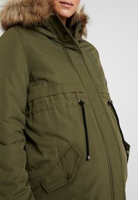 Kvinna i olivgrön vinterjacka med huva prydd med päls, svarta dragskor och framfickor, sedd från bröstet till mitten av låret.