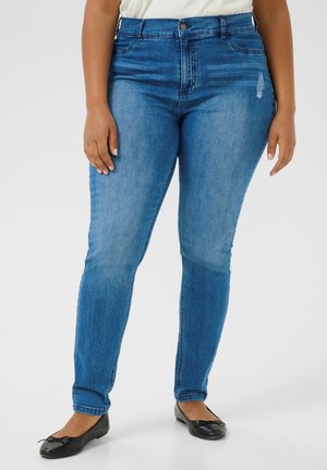 Frau trägt mittelhoch geschnittene blaue Skinny-Jeans mit leichter Abnutzung in der Nähe der Tasche, kombiniert mit schwarzen Ballerinas und einem weißen Top.
