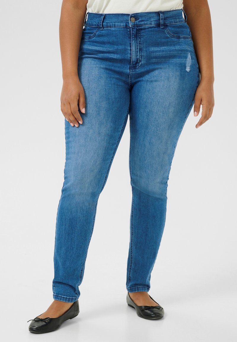 Femme portant un jean skinny bleu taille mi-haute avec une légère usure près de la poche, associé à des ballerines noires et un haut blanc.