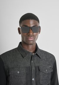 Occhiali da sole con forma piatta e rettangolare e lenti scure. Indossati con una camicia in denim grigia con ampie tasche frontali e chiusure a bottoni.