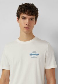 Młody mężczyzna z kręconymi brązowymi włosami, ubrany w gładką białą koszulkę z małym niebieskim logo "S.Oliver Clothing Co." na piersi.