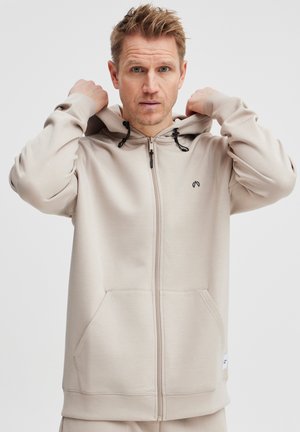 Béžová mikina na zips s kapucňou na stiahnutie, prednými vreckami a malým logom na hrudi. Vyrobená z hladkej, jemnej tkaniny.