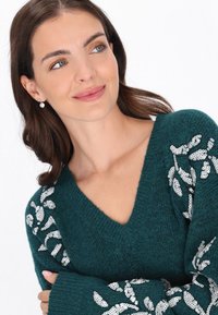 Türkiz kötött pulóver, V-nyakkivágással, ezüst flitteres levélmintákkal az ujjakon. Texturált anyag és laza szabású ujjak.