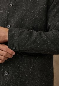 Detaliu al unei jachete negre cu model herringbone, evidențiind țesătura texturată cu ciorchini albi, având manșete cu nasturi și nasturi în față, subliniind designul structurat.