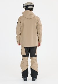 Beige vattentålig jacka med huva, ihopparad med svarta och beiga snöbyxor. Funktioner inkluderar fickor och förstärkta knäområden. Svarta stövlar synliga.