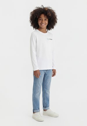 Χαμογελαστό παιδί με σγουρά μαλλιά, που φοράει λευκή μακρυμάνικη μπλούζα με το λογότυπο "MESSI", ανοιχτό μπλε τζιν με γυρισμένα μπατζάκια και λευκά αθλητικά παπούτσια, όρθιο.