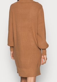 Vue arrière d'une personne portant une robe pull en maille côtelée marron, longue à manches longues et arrivant au genou, avec des manches à revers.