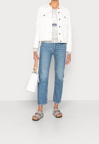 Veste en denim blanc sur un sweat-shirt gris, jean droit bleu, sandales grises avec des nœuds, et un sac blanc avec une poignée tressée.