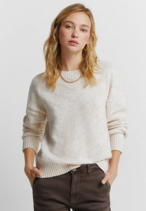 Vaaleanbeige neulepaita, jossa on kohokuvioitu chevron-kuvio, resorit hihoissa ja helmassa. Yhdistetty tummanruskeisiin housuihin. Rento istuvuus.