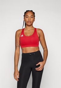 Sostén deportivo rojo con escote en u y tirantes anchos, que presenta un logotipo blanco. Combinado con leggins negros de talle alto y detalle de cordón.