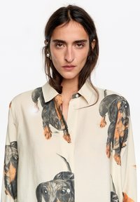 Koszula z długim rękawem z jasnokremowej tkaniny, ozdobiona powtarzającym się nadrukiem jamnika w czerni i pomarańczy, z kołnierzykiem zapinanym na guziki i guzikami z przodu.