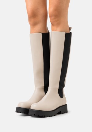 Béžové boty ke kolenům s robustními černými podrážkami a černými elastickými bočními panely, nošené na bosých nohách na neutrálním pozadí.