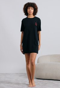 Μαύρο βαμβακερό φόρεμα T-shirt με κοντά μανίκια, διαθέτει ένα μικρό κόκκινο λογότυπο καρδιάς στο στήθος και έχει χαλαρή εφαρμογή, φτάνει πάνω από τα γόνατα.