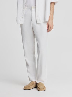 Persona che indossa pantaloni bianchi a gamba larga e una camicia bianca con scarpe slip-on color sabbia, in piedi su uno sfondo chiaro e uniforme.