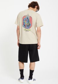Beige graphic t-shirt featuring colorful dragon and wing design on the back, paired with black shorts and white sneakers with blue stripes.