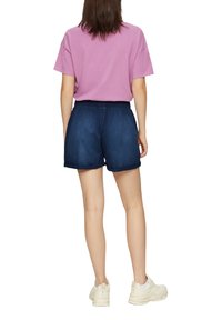 T-shirt en coton violet associé à un short en jean bleu foncé, ample. Le short a une taille élastique et des ourlets pliés, porté avec des baskets blanches.