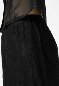 Czarne koronkowe spodnie z elastycznym paskiem, ozdobione delikatnym kwiatowym wzorem i wykonane z miękkiego, lekkiego materiału. Częściowo widoczna prześwitująca czarna bluzka.