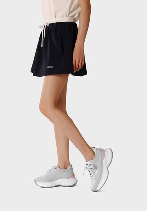 Donna con gonna nera, top bianco e sneakers grigio chiaro, in piedi con un piede leggermente sollevato contro uno sfondo semplice.