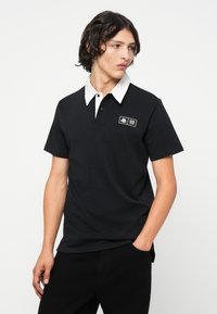 Polo negru cu guler alb, mâneci scurte și o mică ecuson pe partea stângă. Material neted cu o croială clasică.