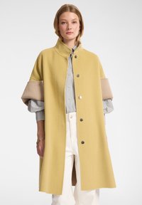 Abrigo largo de lana en amarillo mostaza con un cuello alto y botones plateados. Presenta acentos de tela beige en las mangas. Camisa a rayas debajo.