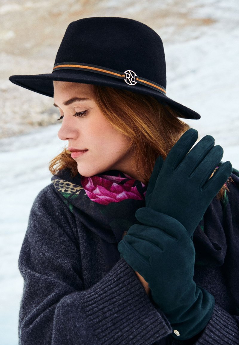 Schwarzer Filzhut mit braunem Band, dunkelgrüne Wildlederhandschuhe und ein florales Tuch, mit einem Nahaufnahme von einer Hand, die das Tuch hält.
