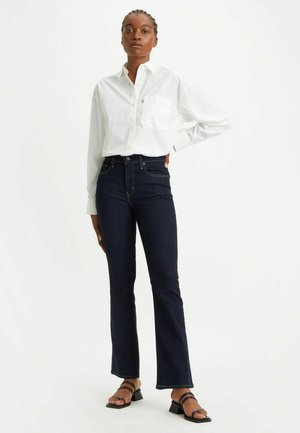 Camisa blanca de cuello, jeans oscuros de cintura alta y sandalias negras de tiras. Los jeans tienen un dobladillo acampanado y detalles de costura sutiles.