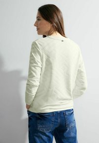 Hellgrünes Langarmshirt mit einem strukturierten geometrischen Muster auf dem Rücken und einem kleinen Logo am Hals. Getragen mit blauen Denim-Jeans.