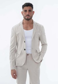 Blazer beige ajusté avec deux boutons noirs, col à revers et poches latérales, associé à un débardeur blanc et un pantalon beige.