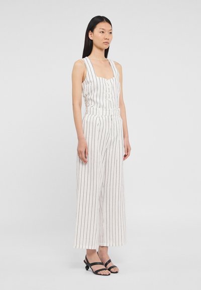 Combinaison blanche avec des rayures noires verticales, décolleté carré, fermeture à boutons à l'avant, jambes larges et ceinture. Portée avec des sandales noires à lanières.
