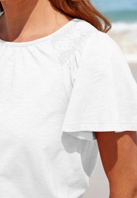 Top blanco de algodón con mangas cortas y voladas, y detalle arrugado en el hombro. La tela parece suave con una textura sutil.