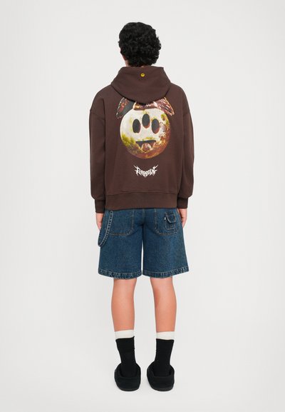 Person with curly hair wearing a brown hoodie with a three-eyed moon graphic and blue denim shorts, standing facing away.