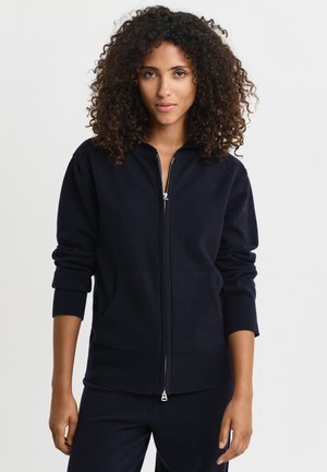 Frau mit lockigem Haar, die eine schwarze Kapuzenjacke mit Reißverschluss und passende schwarze Hose trägt, vor einem schlichten hellen Hintergrund stehend.