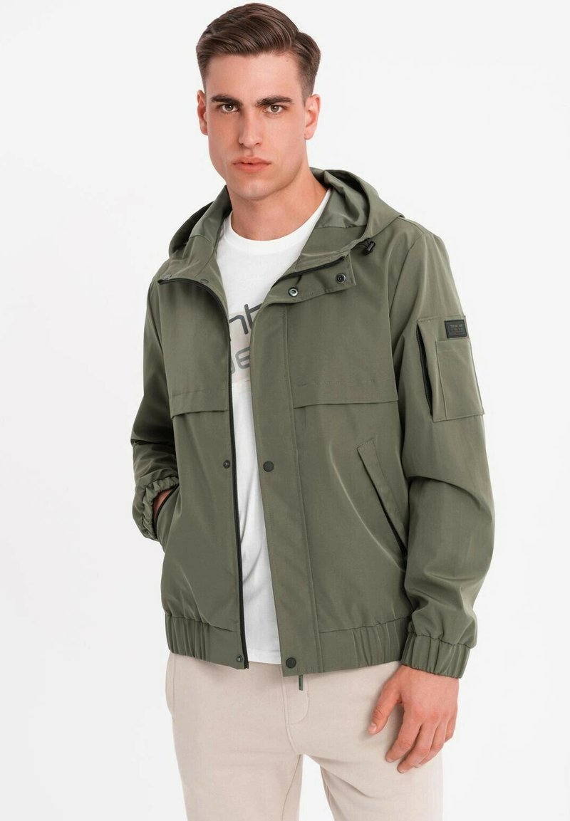 Muž v olivově zelené kapucínové bundě přes bílém tričku s béžovými kalhotami, stojící na jednobarevném bílém pozadí.