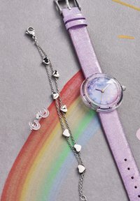 Orologio viola glitter con quadrante rotondo, dettagli argentati e cinturino lavanda; braccialetto a forma di cuore in argento e orecchini a bottone completano il design.