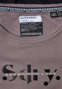 Grå bomullströja med korta ärmar. Har svarttryckt text "Sdy." och "SUPERDRY" på framsidan. Etiketten visar varumärke och skötselråd.