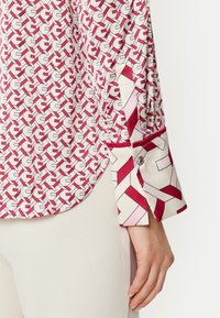 Blusa a righe rosse e bianche con design geometrico, dotata di polsini con bottoni e orlo curvo. Il tessuto ha una consistenza liscia e setosa.