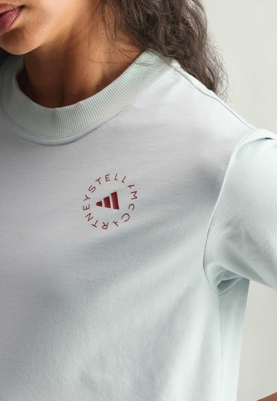 Nahaufnahme einer Person, die ein hellblaues Sweatshirt mit rotem, rundem Stella McCartney-Logo auf der Brust trägt.