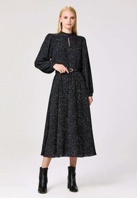 Robe noire à manches longues avec un détail de décolleté en trou de clé, dotée d'une taille ajustée, d'une jupe fluide et de taches blanches éparpillées, associée à des bottines noires.