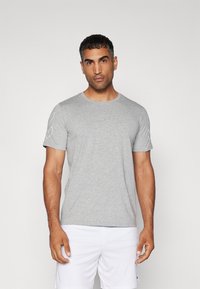Grå bomulds t-shirt med rund halsudskæring og korte ærmer; har et hvidt logodetalje på venstre skulder. Glat tekstur.