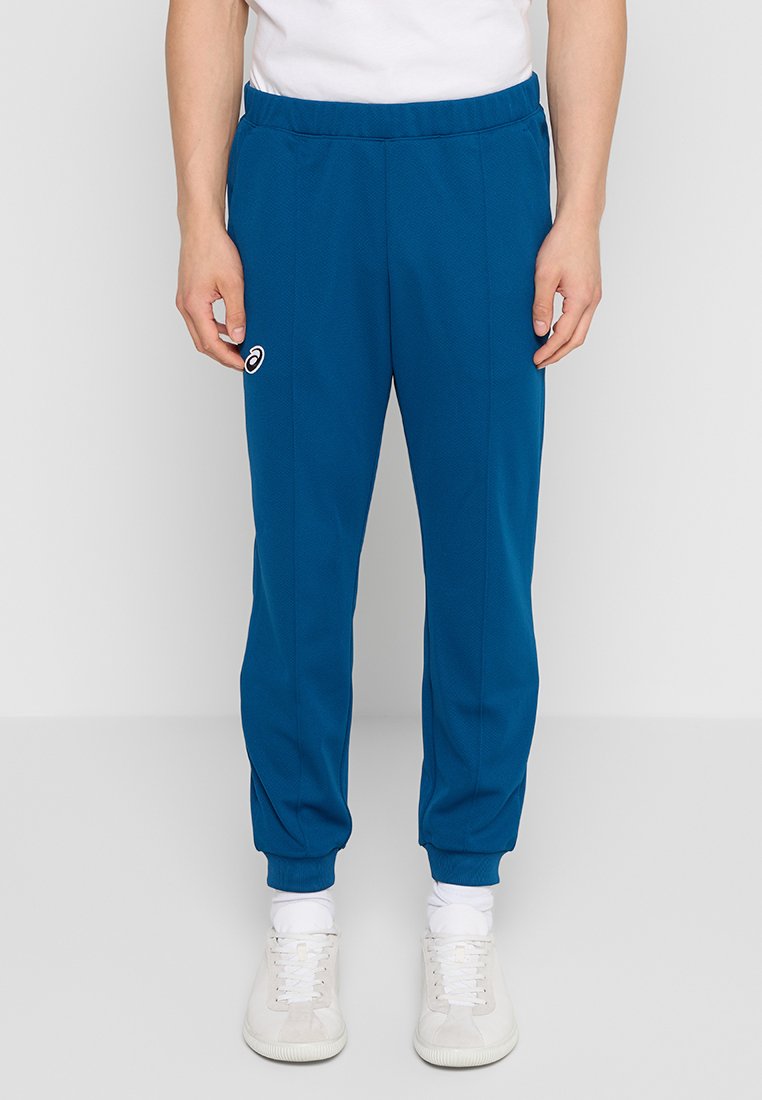Mann in blauen Sportleggings mit elastischen Bündchen und weißen Sneakers, der vor einem schlichten hellgrauen Hintergrund steht.