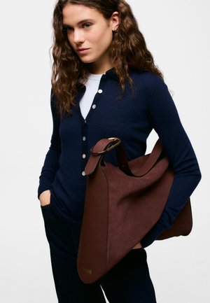 Donna con capelli castani ricci che indossa un cardigan blu navy e una camicia bianca, con una grande borsa a tracolla in pelle marrone con fibbia in ottone.