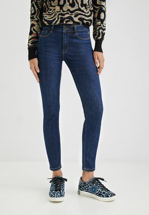 Kvinde iført mørkeblå skinny jeans, farverig trøje med abstrakt mønster og blå sneakers med leopardprint og sorte snørebånd.