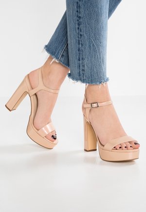 Feet wearing nude patent leather platform high heel sandals with ankle straps and frayed hem blue jeans on white floor.