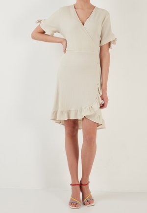 Femme portant une robe beige portefeuille avec un ourlet à volants et des manches courtes nouées aux poignets, associée à des sandales à talons hauts colorées.