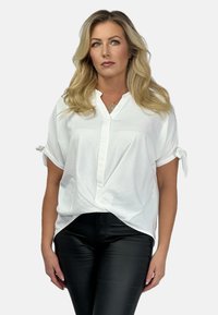 Weiße Bluse mit kurzen Ärmeln, knöpfbarem V-Ausschnitt und geknoteten Ärmeldetails, kombiniert mit schwarzen, schlichten Lederhosen. Glattes, leichtes Material.