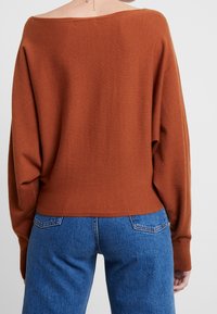 Locker sitzender Pullover mit Carmenausschnitt aus rostfarbenem Strickstoff. Verfügt über herabgesetzte Schultern und gerippte Bündchen. Kombiniert mit blauen Denim-Jeans.