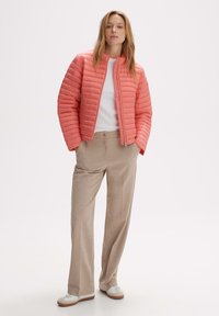 Veste rembourrée corail avec un col haut et des coutures horizontales, associée à un pantalon large beige et des baskets blanches.