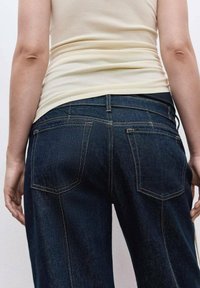 Person med en beige ärmlös topp och mörkblå denimjeans, visad bakifrån med armarna avslappnat vid sidorna.